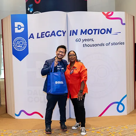 Two Dallas College employees pose in front of an oversized book sculpture that reads: A LEGACY IN MOTION, 60 years, thousands of stories.