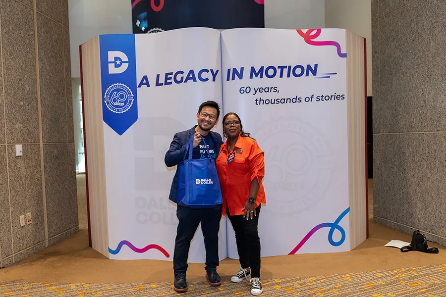 Two Dallas College employees pose in front of an oversized book sculpture that reads: A LEGACY IN MOTION, 60 years, thousands of stories.