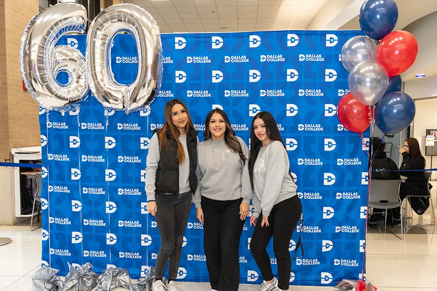 Three students pose in front of Dallas College backdrop and balloons.