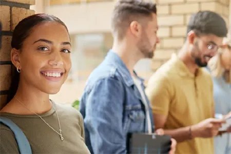 Group of diverse college students standing in a campus hallway with backpacks and textbooks; one student smiles at the camera while others read or talk nearby.