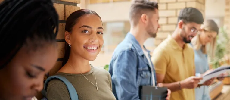 Group of diverse college students standing in a campus hallway with backpacks and textbooks; one student smiles at the camera while others read or talk nearby.