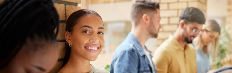 Group of diverse college students standing in a campus hallway with backpacks and textbooks; one student smiles at the camera while others read or talk nearby.