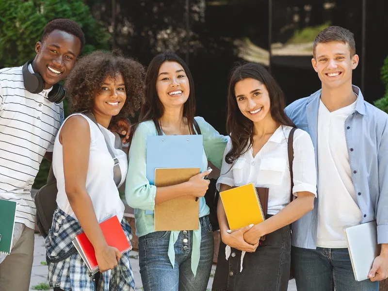 Five students standing outdoors with notebooks, greenery and building in background.