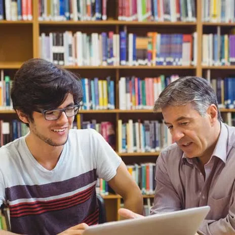 A student and teacher engage with a laptop in a library, surrounded by bookshelves
