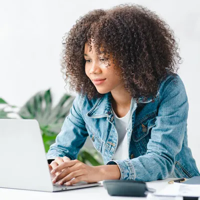 A woman with curly hair in a denim jacket focused on a laptop at a white desk, surrounded by plants, conveying a calm and productive atmosphere