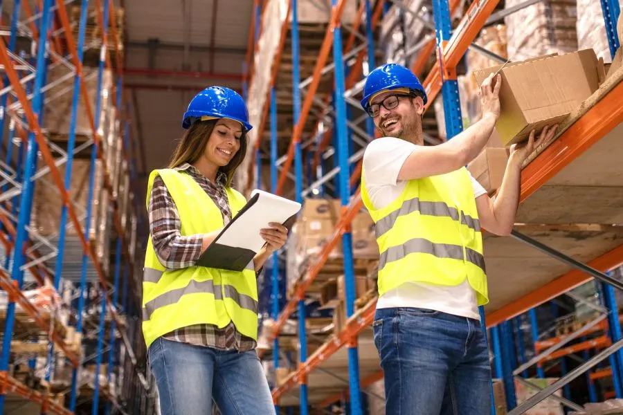 Two workers review inventory in a warehouse. One checks the list while the other reaches for a box on the shelf.