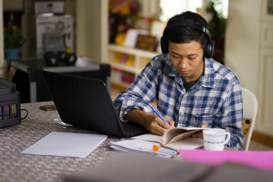 A student sits at a kitchen table and studies with their laptop, notebooks and headphones on