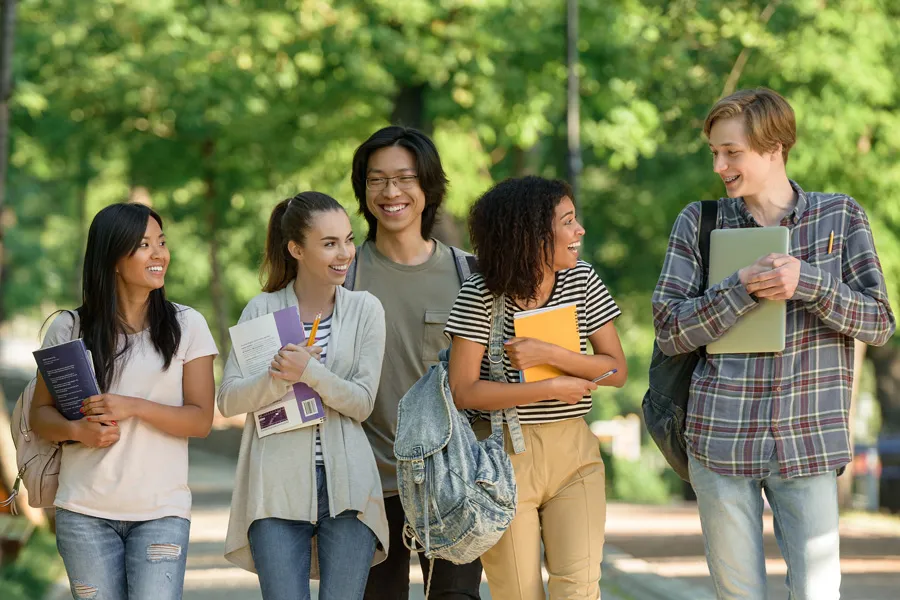 College students walking on a campus pathway surrounded by greenery, carrying books and backpacks