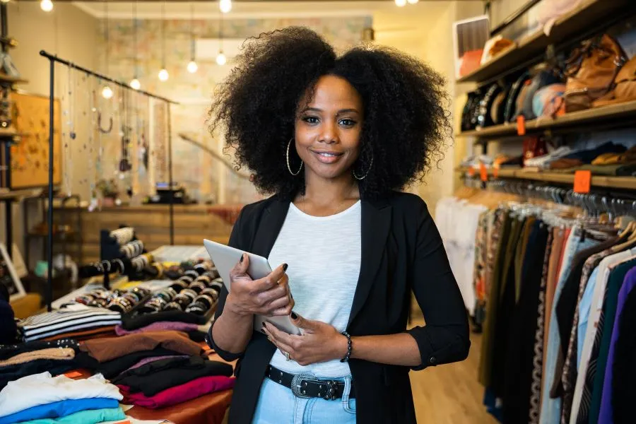 A clothing store owner stands with a tablet in her hands in front of shelves and racks of clothes
