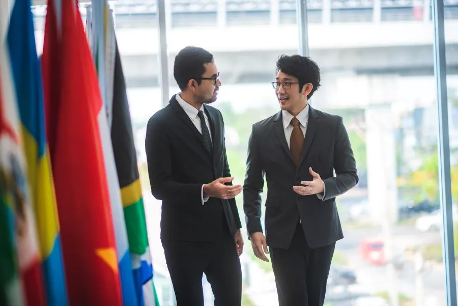 Two men wearing suits talk in front of a wall of international flags