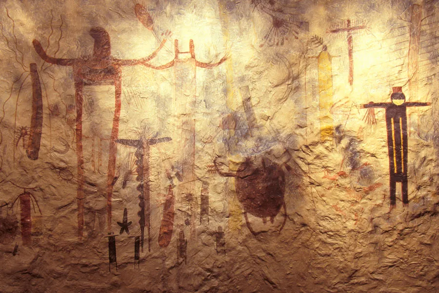 Ancient cave paintings on a textured stone surface, featuring human-like figures and abstract symbols in red, black, and yellow hues.