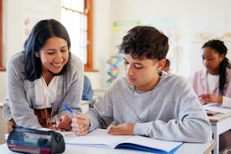 eacher assisting a student at a desk with an open notebook in a classroom; another student is seated in the background.