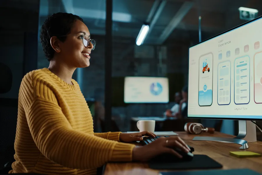 Person working at a desk on a large monitor displaying mobile app user interface designs, with a keyboard, mouse, coffee mug, and headphones on the desk in a modern office setting.
