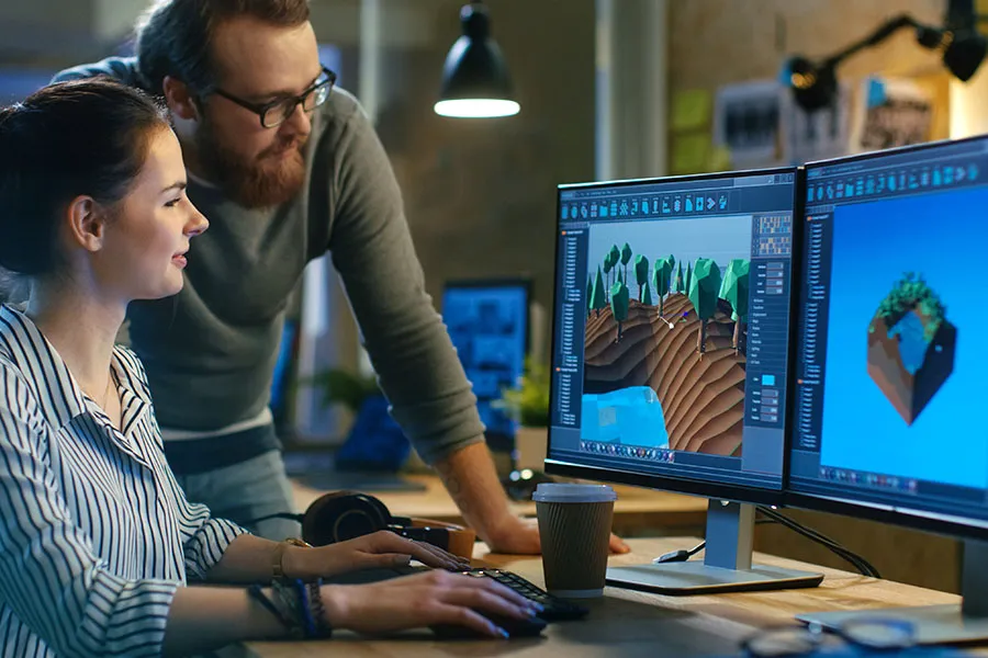 Two people collaborate in an office, using 3D modeling software on dual monitors; one is seated, the other is standing.