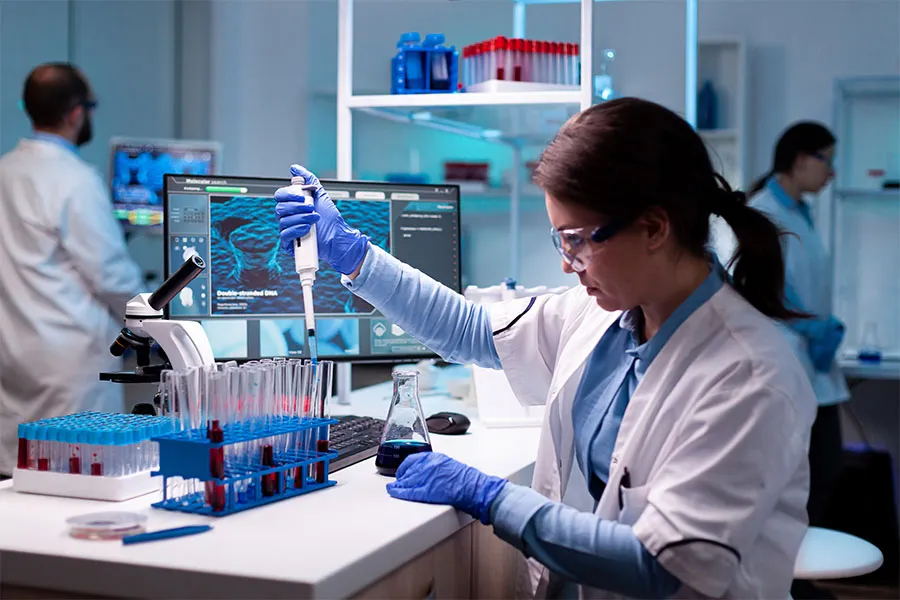 Scientist in white lab coat and blue gloves uses a pipette with test tubes; two others work in the background with lab equipment.
