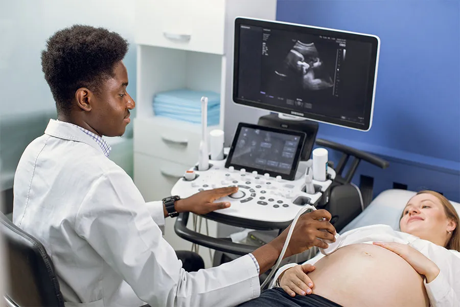 Medical professional performs an ultrasound on a pregnant patient; fetus image is visible on the monitor, with faces blurred for privacy.