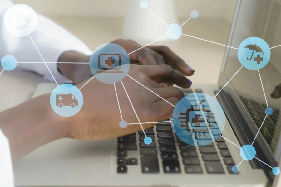 Hands typing on a laptop keyboard with healthcare icons like a cross, ambulance, and stethoscope connected by lines.