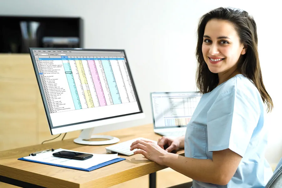 Person in a light blue shirt works at a wooden desk with a computer and laptop displaying spreadsheets.