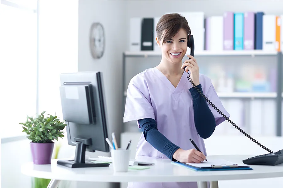Person in scrubs stands at a medical front office desk, talking on the phone and writing on a clipboard; desk has a monitor and supplies.
