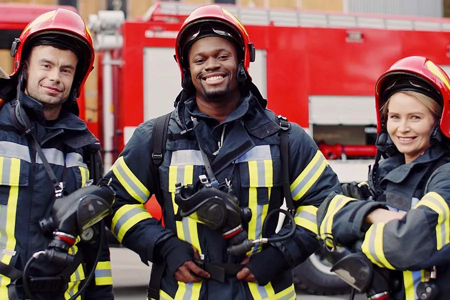 Three firefighters in helmets and reflective jackets stand in front of a red fire truck.