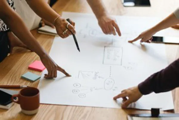 People gather around a table and point to various areas of a flowchart spread out on a table