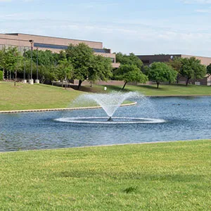 A vibrant campus scene with a large fountain in a pond, surrounded by lush green grass and trees, and modern brick buildings in the background under a blue sky