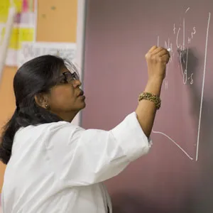 A woman in a white lab coat writes scientific equations on a chalkboard