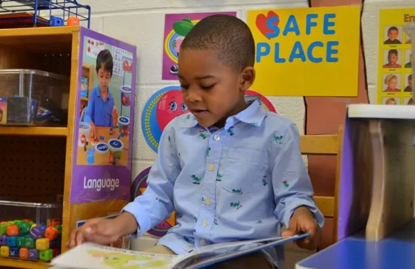Child reading a book in a classroom with educational posters and toys in the background.