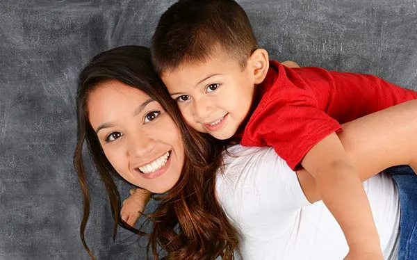 Woman with long hair carrying a child on their back against a gray textured background.