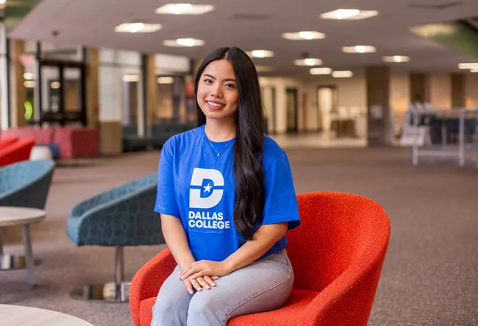 female student wearing Dallas College shirt sitting in chair and smiling