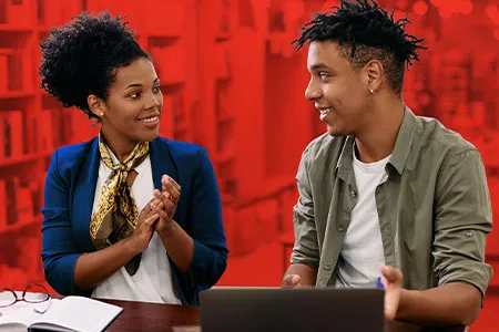 a young man and a young woman talk while sitting at a table in the library
