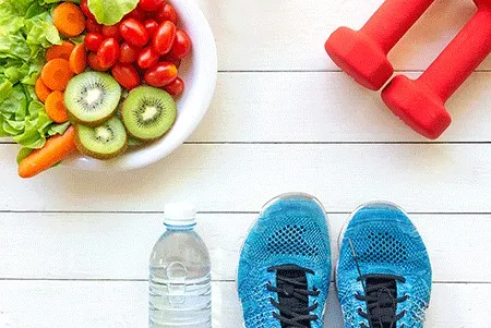 a bowl with fruits and vegetables, hand weights, water bottle and athletic shoes