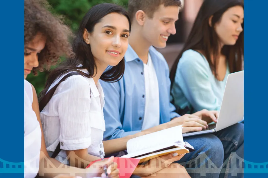 a row of students sitting outside, working on laptops or notebooks