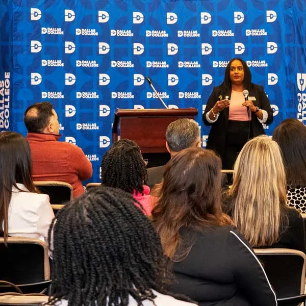 Audience seated in rows listening to a speaker standing at a podium in front of a Dallas College backdrop.