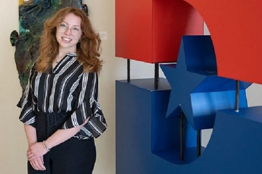 Ava Stipe in formal attire in a gallery, posing and smiling next to a Dallas College logo.