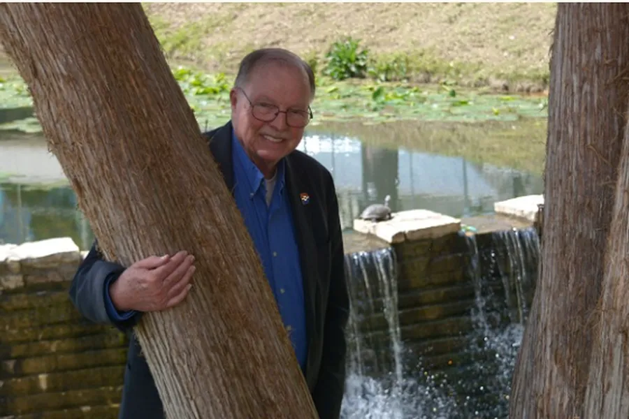 Dr. Paul Benson wearing a black blazer with a Dallas college logo on the shoulder of his blazer has his arm wrapped along a tree in front of moving water overflowing a short brick ponywall.