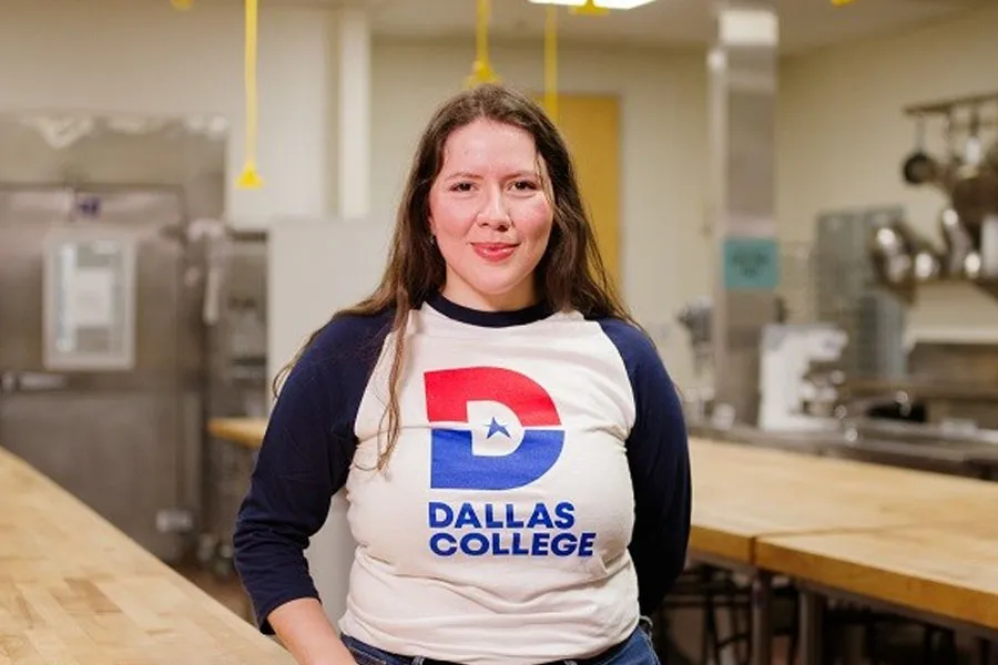 Frida Hutchinson standing and smiling at the camera, wearing a Dallas College shirt. A kitchen background is visible.
