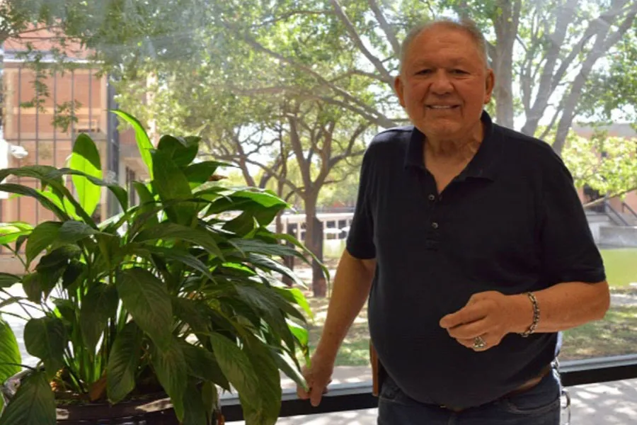 Jim Stover smiles at the camera on a Dallas College campus, where nature and buildings are visible in the background.