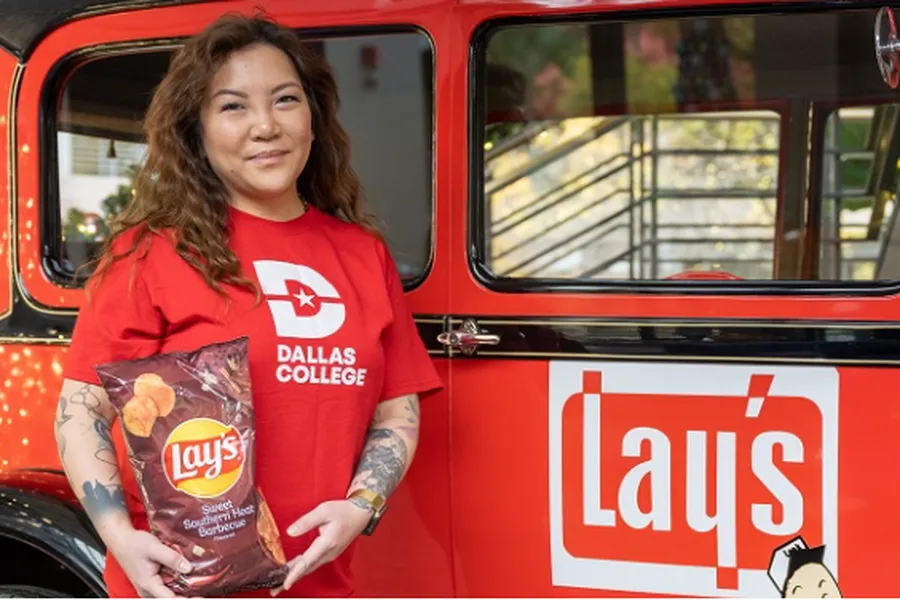 Ngoc Trinh in a red shirt with the Dallas Logo logo on it holds a bag of Lay's sweet southern heat barbecue chips while standing in front of a red vehicle with the Lays logo on it.