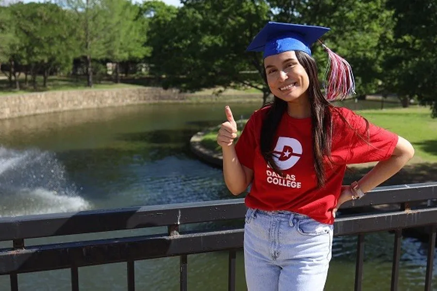 Cano Padron wearing a red Dallas College shirt and a graduation cap. He points a thumb-up and smiles at the camera, with a visible fountain and field in the background.