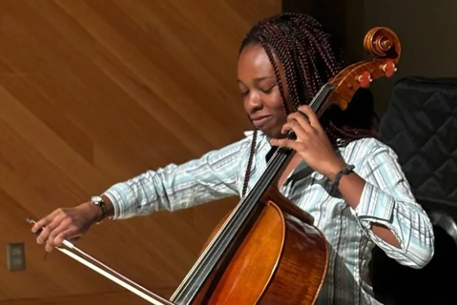 Skylar Cambell plays the Cello in front of a piano with a quilt like cover on it and a wall with a wooden design..