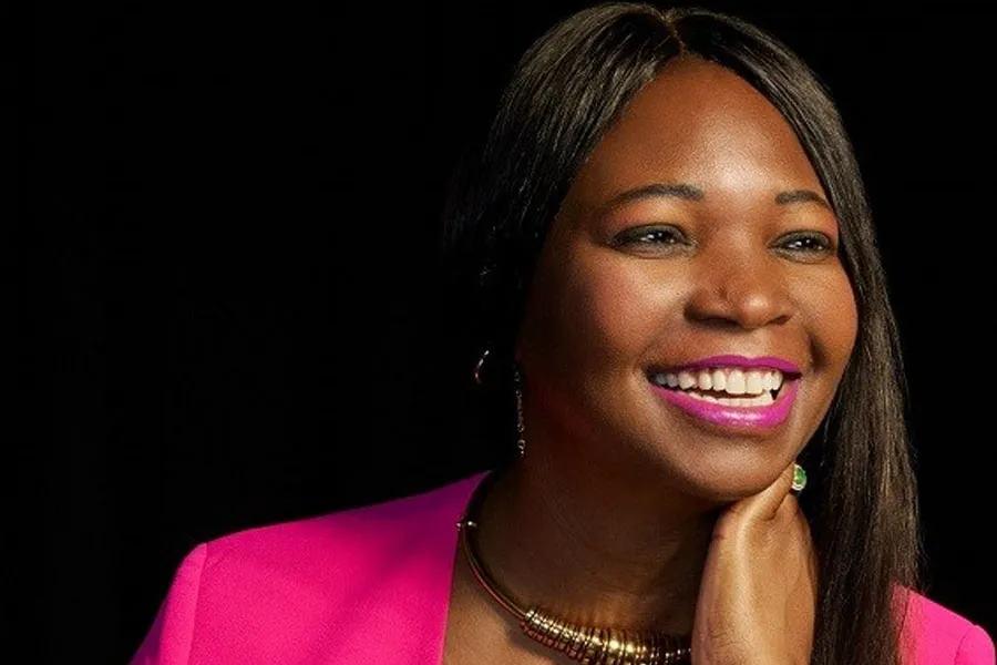 Ebbah Realzola smiling, facing away from the camera. She wears a hot-pink blazer and has her left hand at her neck. An all-black background is visible.