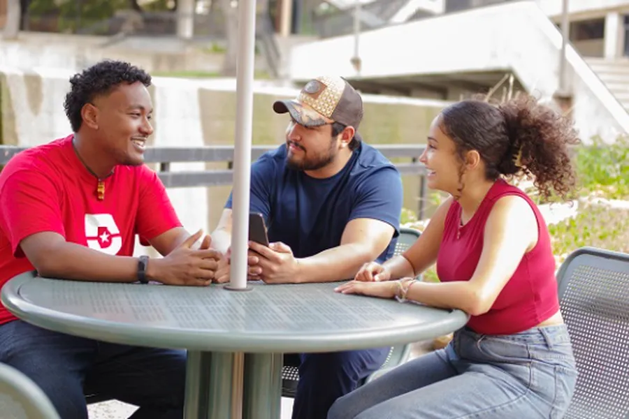 Yonata Kidane wearing a red shirt with the Dallas College logo talks with 2 other students sitting at a circular, metal picnic table.