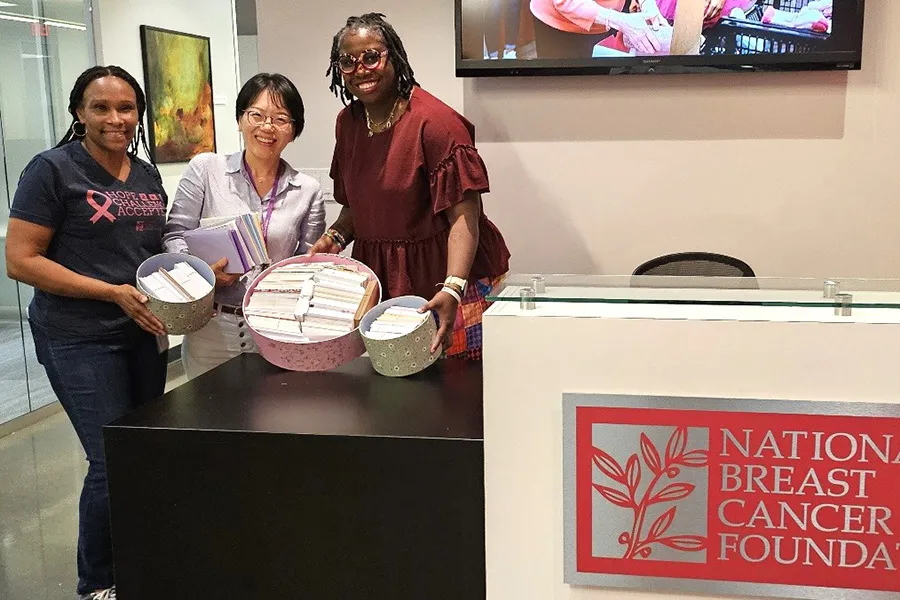 A group of smiling individuals stand together indoors holding stacks of colorful handwritten encouragement cards, symbolizing Dallas College students’ effort to create and donate 1,000 heartfelt messages of support for breast cancer patients through the National Breast Cancer Foundation