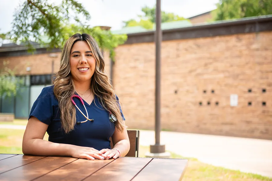 Faviola Cecena in blue scrubs wearing a stethoscope around her neck is sitting at a brown, circular table.