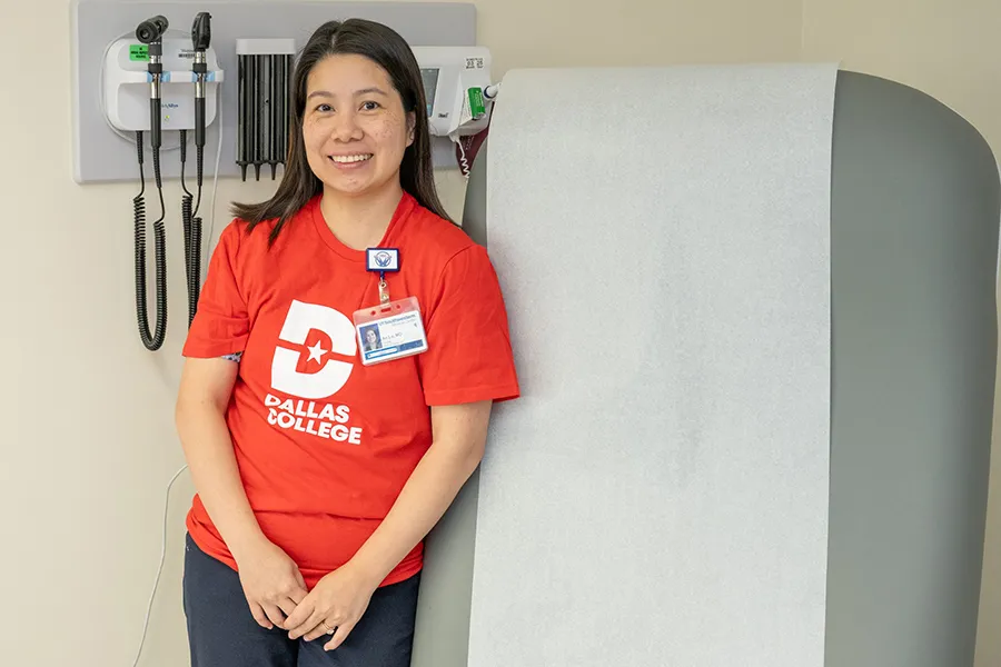 Dr. An Lu standing in front of wall-mounted medical equipment and wearing a Dallas College t-shirt.