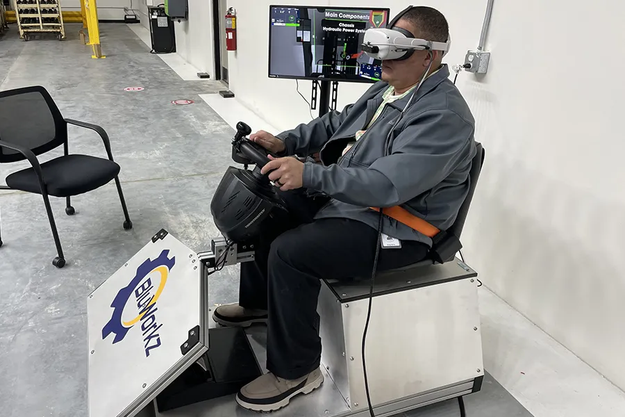 A man is using a forklift simulator that is white with the Blu-Workz logo on the outside. The simulator has a car steering wheel like set up.