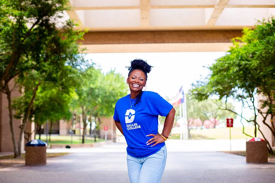 Kristen Bradford in a blue Dallas College shirt stands on a walkway lined with trees.