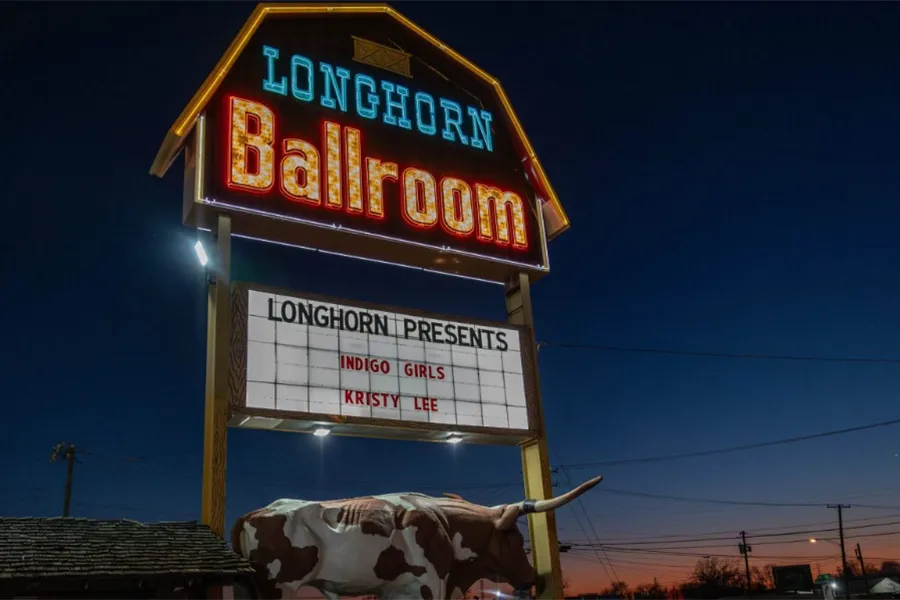 The illuminated Longhorn Ballroom sign stands outside the Dallas music venue at dusk. A marquee beneath the sign advertises upcoming performers, while a longhorn sculpture sits at the base of the sign.