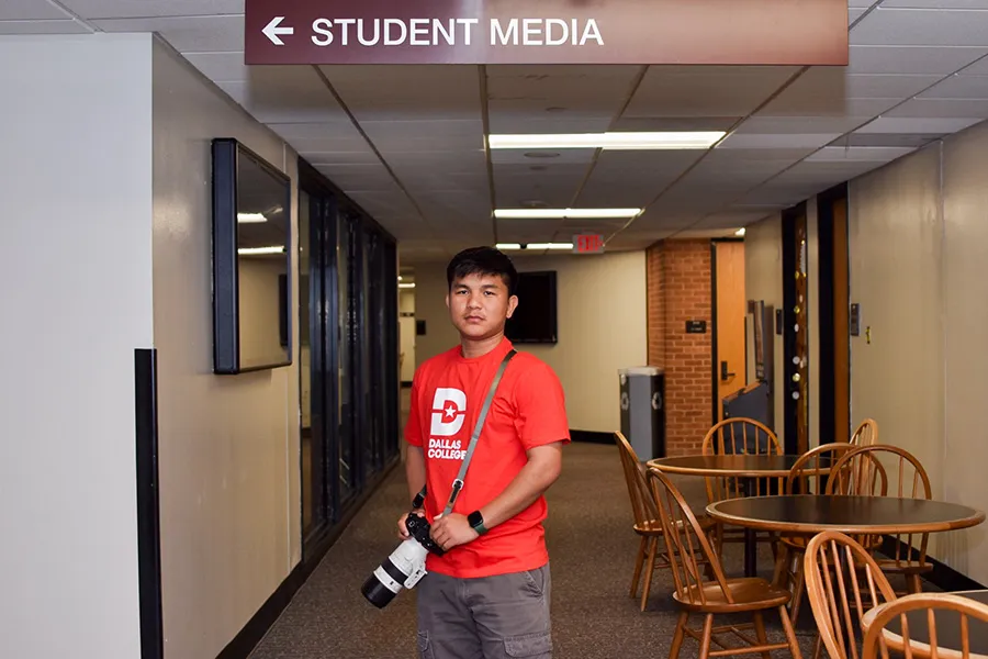 Ning wearing a red Dallas College shirt with a camera strapped around his neck. He holds the camera at a waist-level with two hands as he poses for the picture in a building with a huge sign stating, 